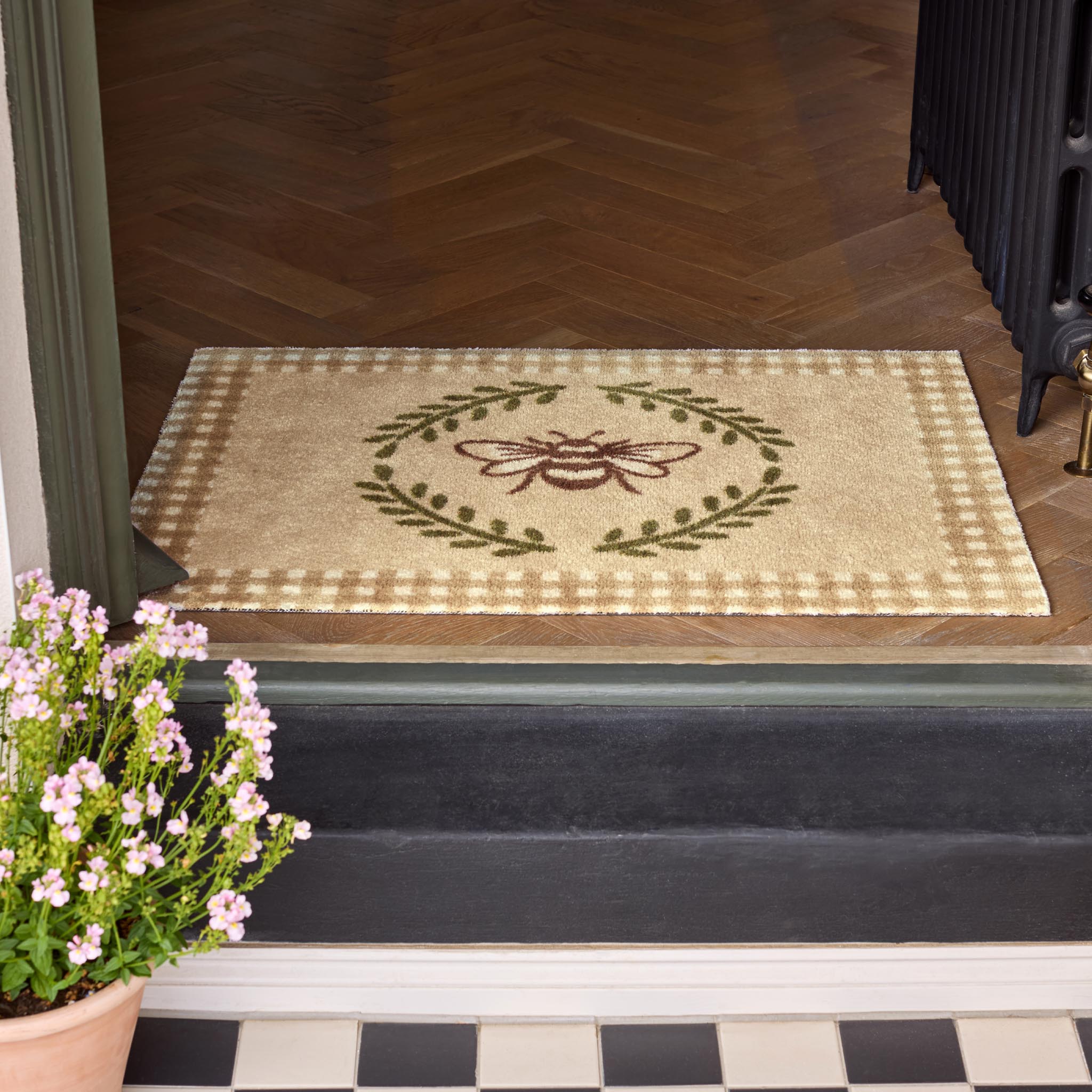 Doormat with bee design on a wooden floor, potted plant to the side.