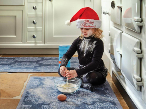 A young girl sat on a kitchen runner cracking an egg into a bowl wearing a santa hat