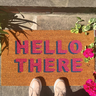 A pair of feet standing on a coir outdoor mat that says hello there in pink writing