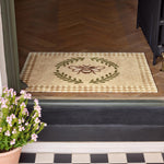 Doormat with bee design on a wooden floor, potted plant to the side.