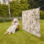 Woman sitting in a chair reading a book next to a floral rug hanging on a washing line in a garden.
